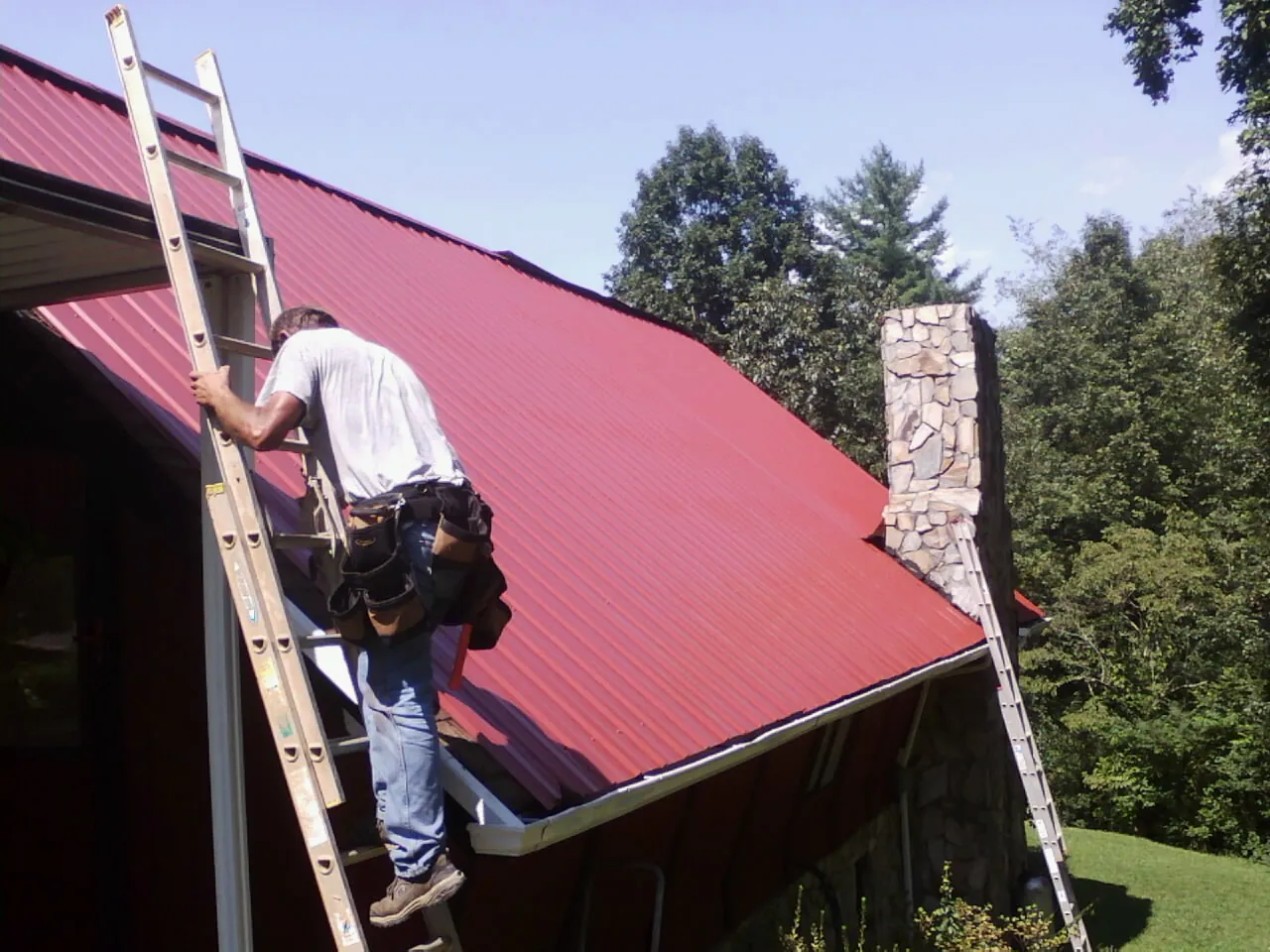 Roofer on a ladder beside a red metal roof and stone chimney — Soffit & Fascia Repair by Roofing Brothers in Midway City